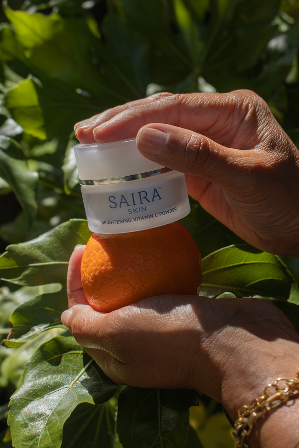 A person holds a jar of SAIRA Brightening Vitamin C in front of a bright orange on a leafy green plant. The hands are in focus, with the background foliage blurred.