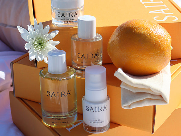 Four SAIRA skincare products arranged in a styled flat-lay with orange packaging, a white flower, and an orange on a folded cream muslin cloth.